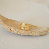 An 18K gold plated stainless steel asymmetric ring with a textured surface, displayed on a natural stone surface.