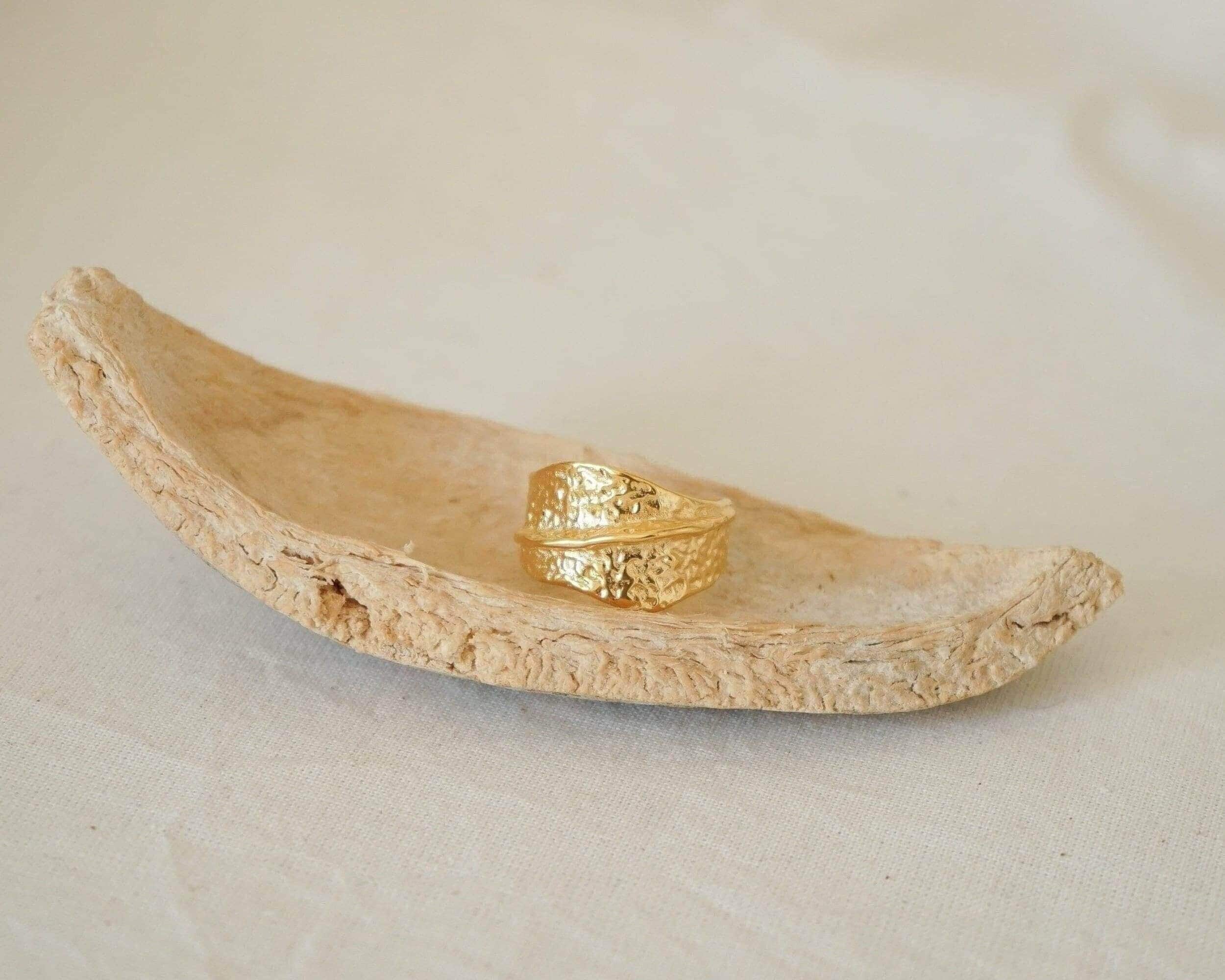 An 18K gold plated stainless steel asymmetric ring with a textured surface, displayed on a natural stone surface.