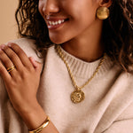Woman wearing large gold disc earrings and achunky gold pendant with a neutral background