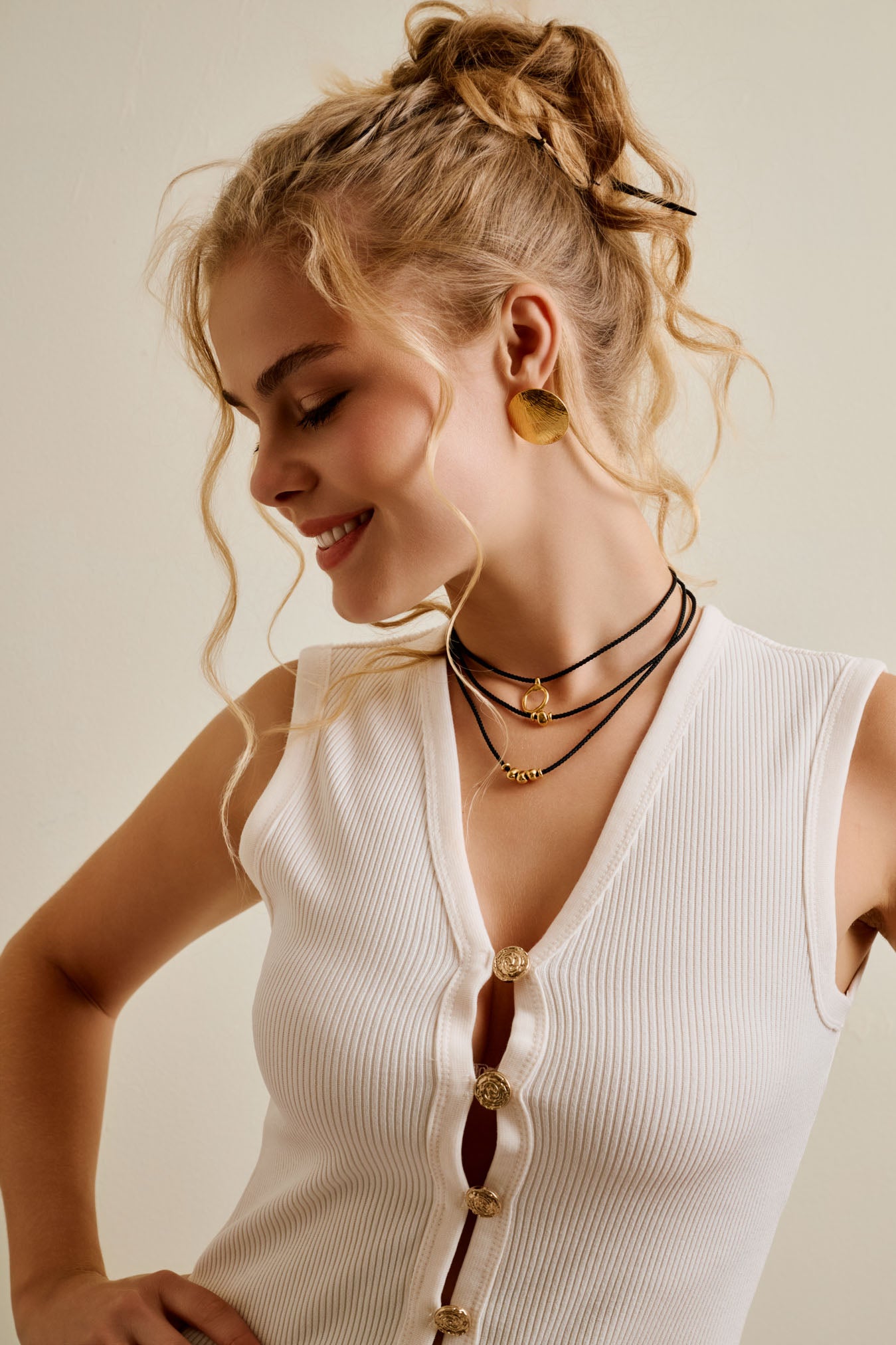 Woman wearing a white sleeveless top with gold buttons and necklaces, against a plain background